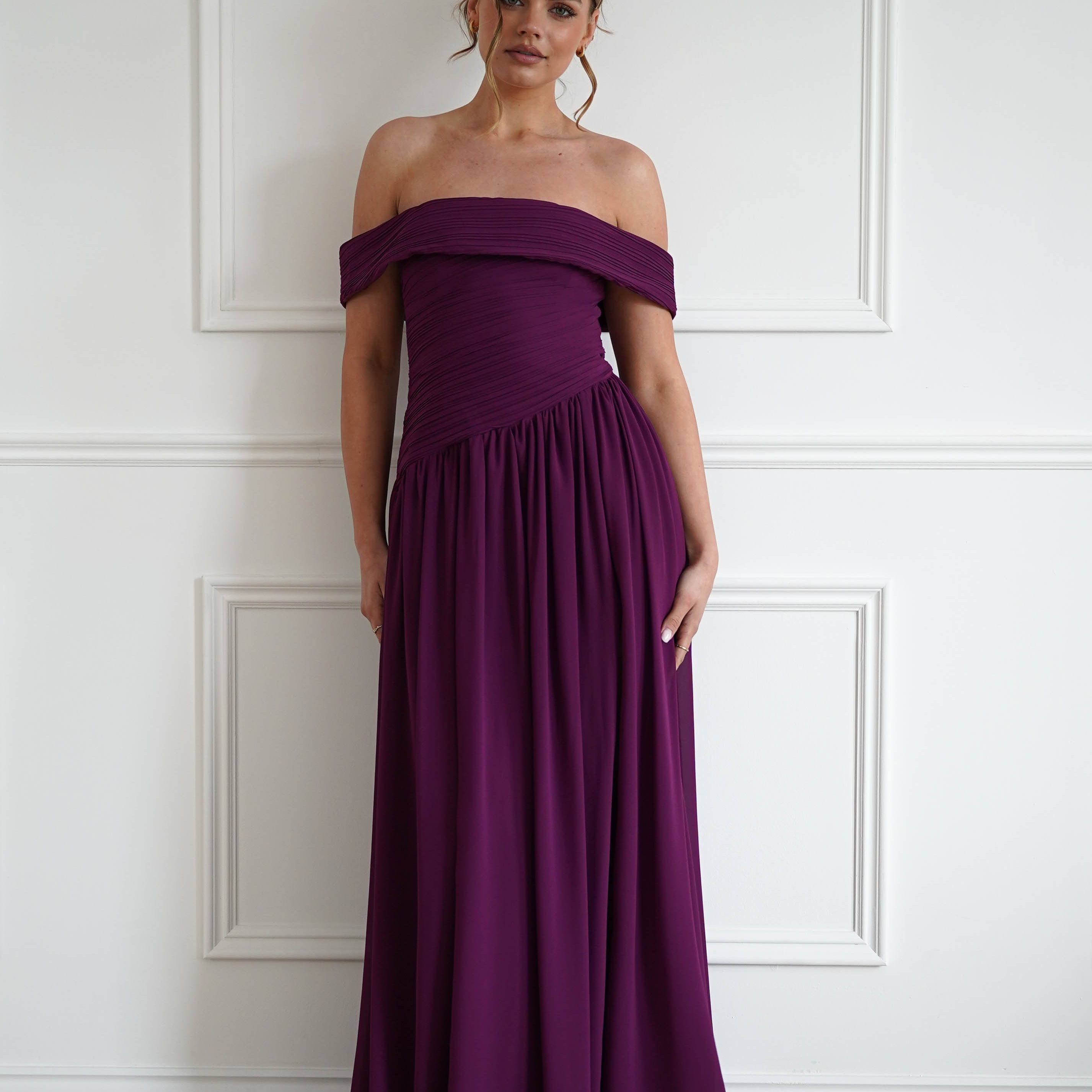 Woman wearing a plum off-the-shoulder chiffon maxi dress with a ruched bodice, gathered waist, and flowing floor-length skirt, styled for weddings, bridesmaid events, and formal occasions.