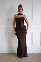Woman wearing a chocolate brown strapless maxi dress with a draped neckline, ruched hip detailing, matching neck scarf, and fitted floor-length silhouette, ideal for weddings, black-tie events, galas, and formal evening occasions.