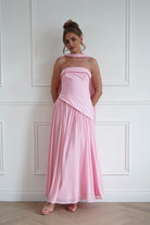 Woman wearing a pink strapless maxi dress with an asymmetric waistline, flowing chiffon skirt, folded neckline detail, and matching scarf accent, perfect for weddings, bridesmaid celebrations, garden parties, and formal occasions.